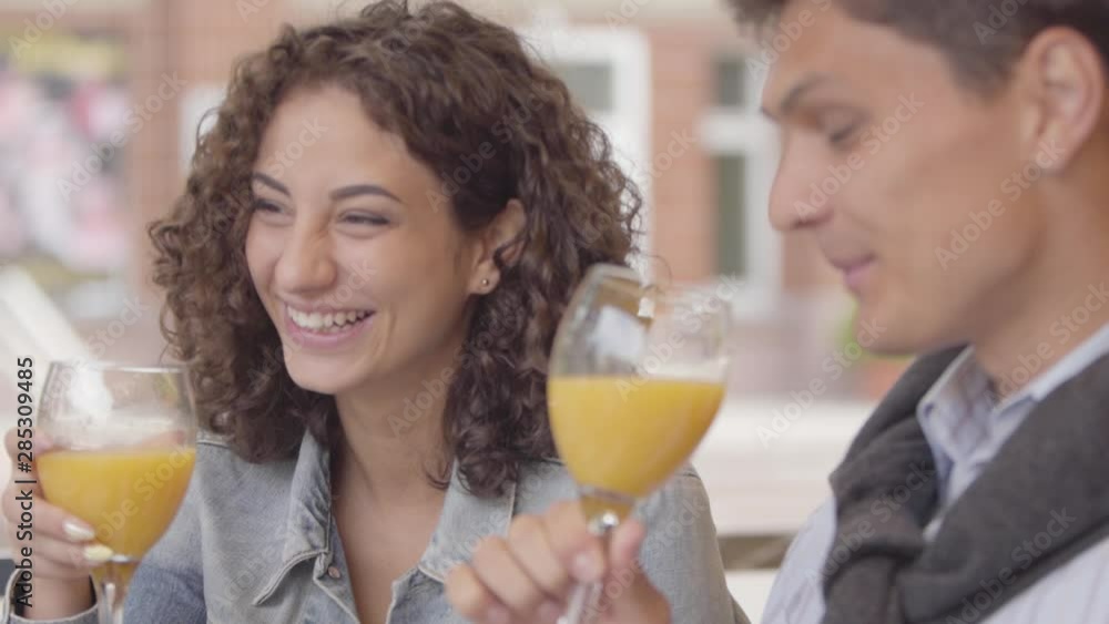 Portrait of a happy couple chatting and laughing while drinking orange juice sitting in cafe outdoors. Young man and woman spending time together enjoying their meal. Date concept. Slow motion.
