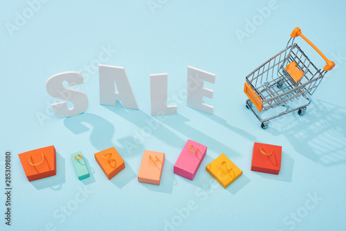 top view of white sale lettering near shopping trolley and colorful shopping bags on blue background