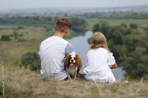 Boy and girl sits on the hill above the river with their smiling spaniel dog