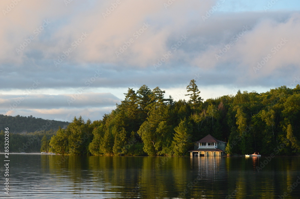 Fototapeta premium Boat house on Saranac Lake bathed in the morning sunrise. The coastline is reflected in the water and framed by the colored clouds