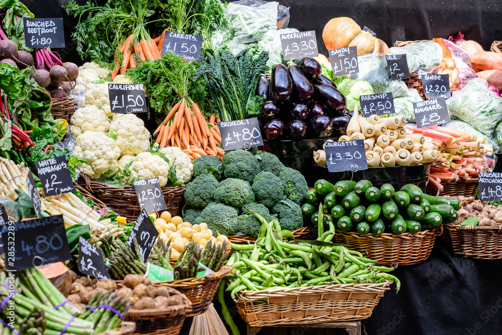 Green organic vegetables in display for sale at a street food market, with names displayed in English, carrots, white asparagus, courgette, aubergine, broccoli, British rhubarb, potatoes, cauliflower,