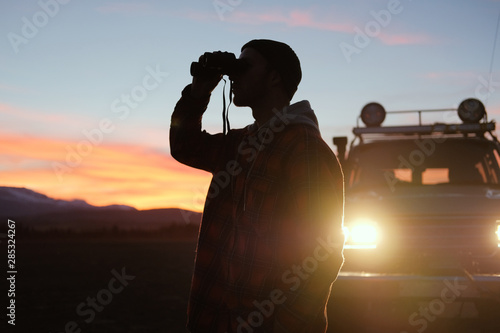 A man with binoculars near the car at sunset. Man Binoculars Looking Mountain Cloudscape Traveling Concept photo
