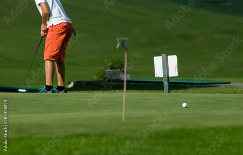 young boy exercising on a green of golf course club to pull ball into the hole, grass, sunset, lesson, sports, concentration, hobby, child, vacation, mountain, Trentino, Alto Adige, Italy