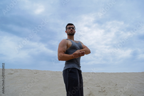 A young muscular man in glasses in a t-shirt stands in the desert against the sky.