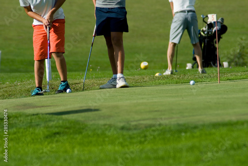 two young boys exercising on a green of golf course to pull the ball into hole, grass, sunset, lesson, sports, concentration, hobby, child, vacation, Trentino, Alto Adige, Italy