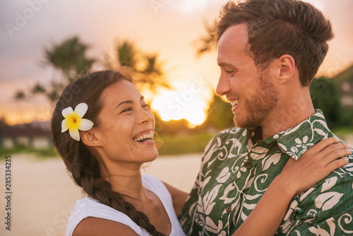 Happy couple lovers in love on romantic sunset beach vacation in Hawaii travel. Asian woman hugging Caucasian man smiling, interracial relationship. Newlyweds honeymoon or wedding on Tahiti beach.
