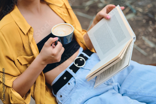 Close up woman in yellow shirt and jeans holding cup of coffee and book in hands in park