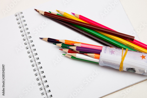 colored pencils wrapped in euro note lie on a white notebook on a white table bright copy space