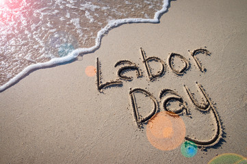Labor Day message handwritten on the smooth sand of an empty beach with sunny lens flare across an oncoming wave