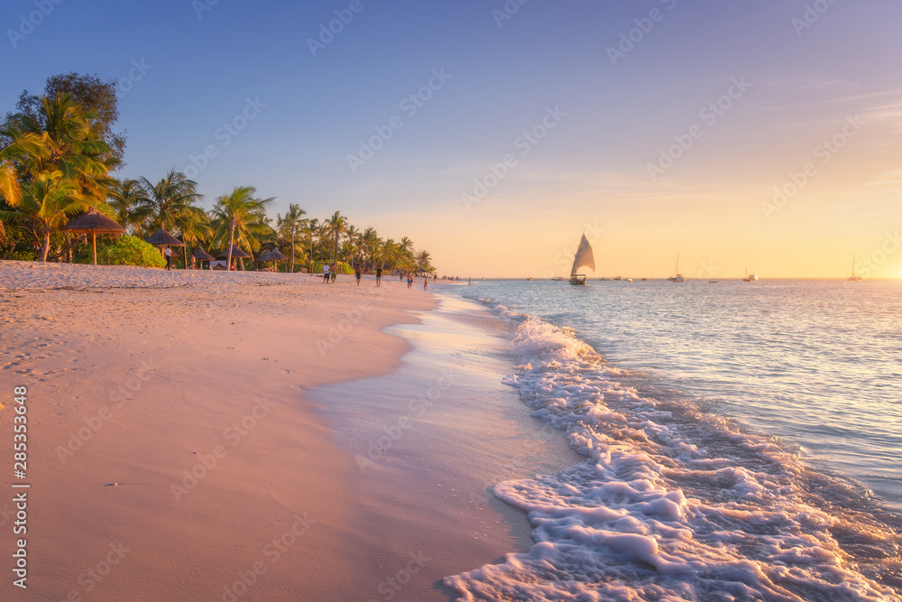 Sandy beach with sea waves, palm trees and walking people at colorful sunset in summer.  Tropical landscape with blue sea, palms, boats and yachts in ocean, beautiful sky. Travel in exotic Africa - obrazy, fototapety, plakaty 