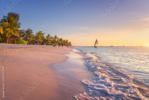 Fototapeta Naklejka Na Ścianę i Meble -  Sandy beach with sea waves, palm trees and walking people at colorful sunset in summer.  Tropical landscape with blue sea, palms, boats and yachts in ocean, beautiful sky. Travel in exotic Africa