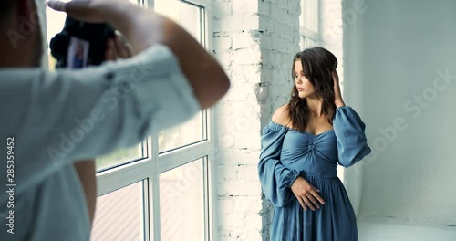 Beautiful caucasian female model in romantic blue dress standing near window and posing photographer, studio photosession