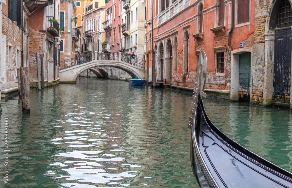 The bow of a gondola, depicting no gondolier, navigating one canal at Venice