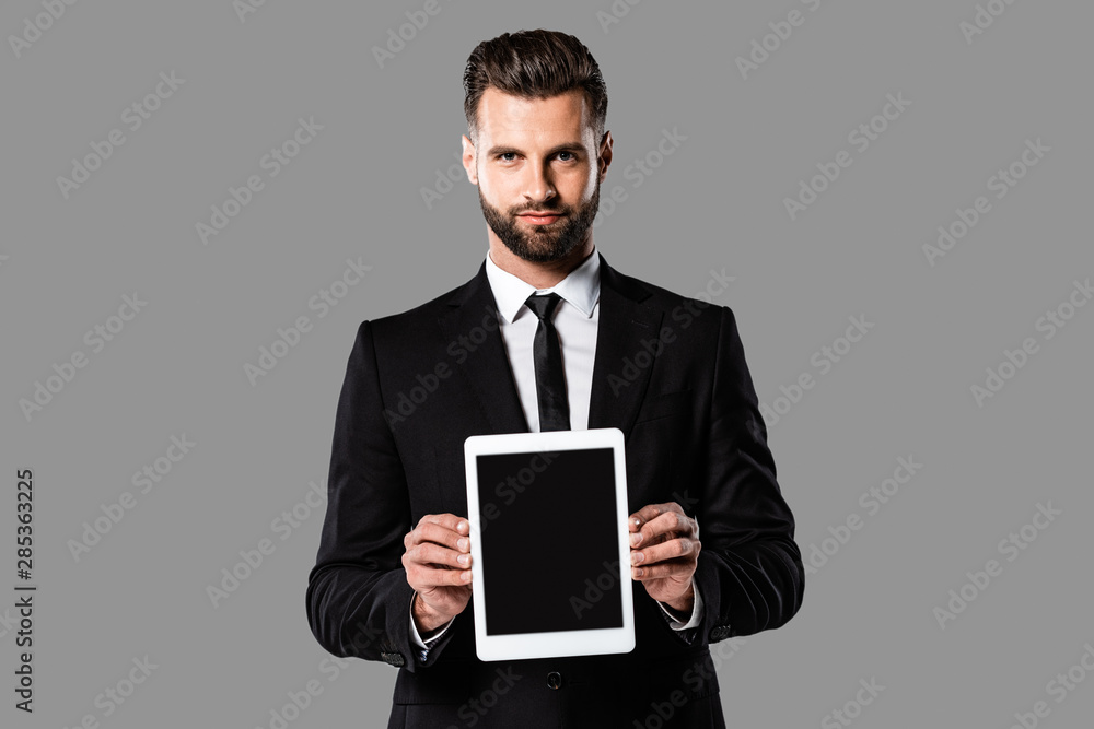 handsome businessman in black suit showing digital tablet with blank screen isolated on grey