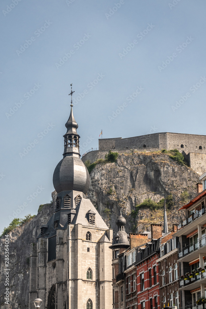 Dinant, Belgium - June 26, 2019: The citadel fort on cliff towers high above the spire of gray stone Notre Dame church under light blue sky. Housing builidngs in differnt colors.