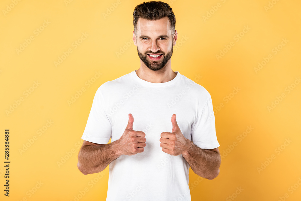 smiling handsome man in white t-shirt looking at camera and showing thumbs up isolated on yellow smiling handsome man in white t-shirt looking at camera and showing thumbs up isolated on yellow
