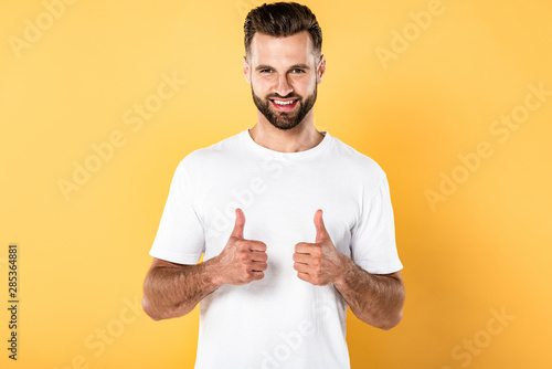 smiling handsome man in white t-shirt looking at camera and showing thumbs up isolated on yellow