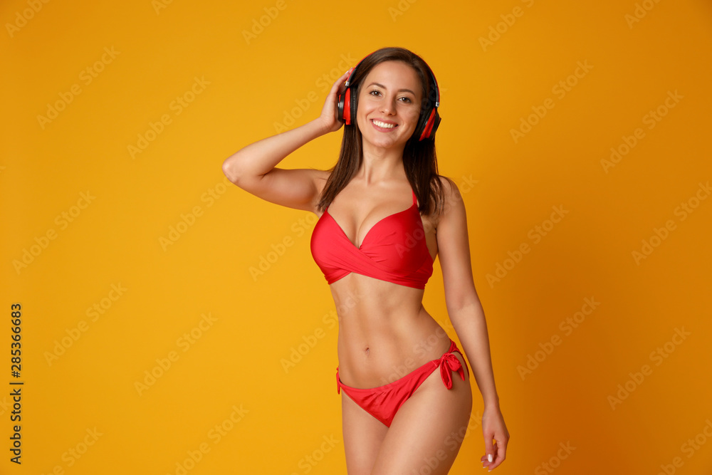 Pretty sexy woman with slim body in stylish red bikini and headphones on orange background, space for text - obrazy, fototapety, plakaty 