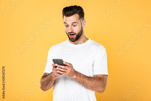 excited handsome man in white t-shirt using smartphone isolated on yellow