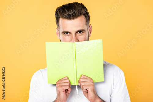 handsome man in white t-shirt holding book in front of face isolated on yellow