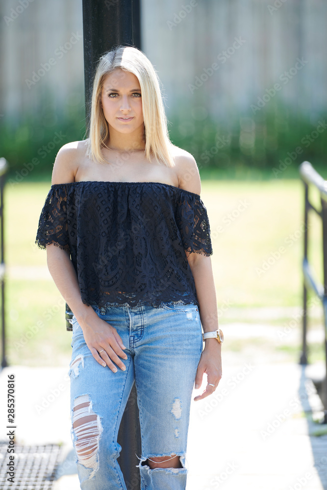 Beautiful young blonde woman poses outside in denim and black off the shoulder blouse