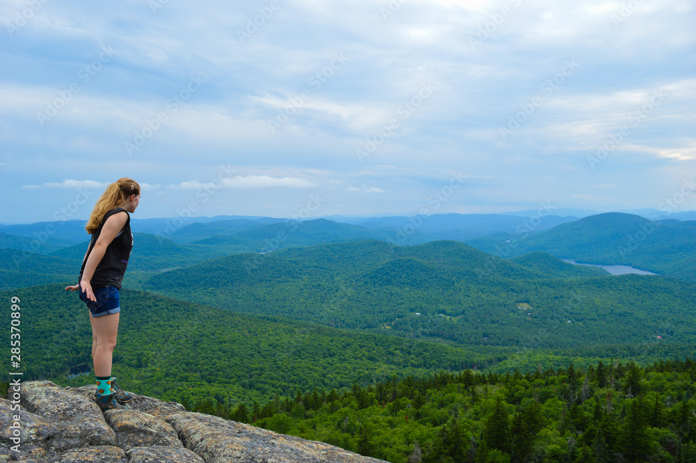 A female hiker cautiously leans over the edge of a mountain, looking down with the Adirondack mountain ranges on display in the distance.