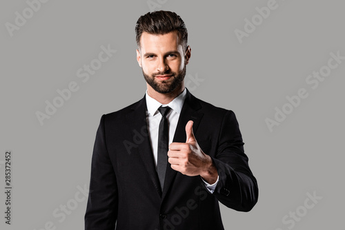smiling handsome businessman in black suit showing thumb up isolated on grey