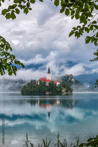 Bled, Slovenia - Beautiful Pilgrimage Church of the Assumption of Maria on Lake Bled (Blejsko Jezero) covered by fog with Bled Castle and Julian Alps on a misty summer morning with foliage frame