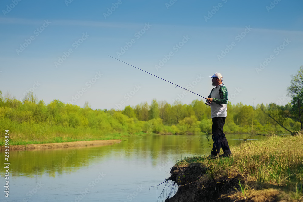 Fisherman with a spinning rod catching fish on a river at sunny summer day with green trees at background. Outdoor weekend activity. Photo with shallow depth of field taken at wide open aperture. Fisherman with a spinning rod catching fish on a river at sunny summer day with green trees at background. Outdoor weekend activity. Photo with shallow depth of field taken at wide open aperture.