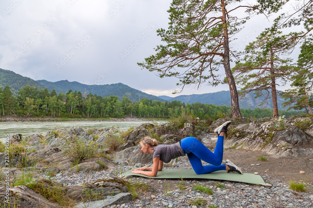 Slim young healthy sports girl doing the donkey kick exercise on all fours arching back straightening leg up concept sport, fitness, lifestyle. Work out and warm up outdoors in mountains near river. Slim young healthy sports girl doing the donkey kick exercise on all fours arching back straightening leg up concept sport, fitness, lifestyle. Work out and warm up outdoors in mountains near river.
