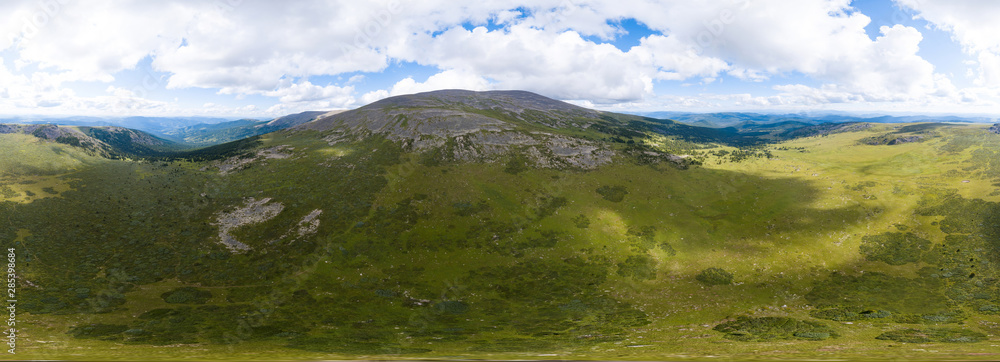 360 degree panoramic aerial view in the wild with green fields and grass and a high mountain with a rocky peak on a warm summer day with white clouds and blue sky with high detail. 3d tour