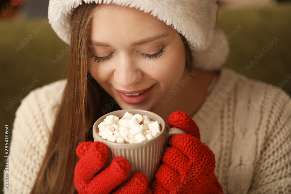 Beautiful woman in Santa Claus hat drinking hot chocolate at home, closeup Beautiful woman in Santa Claus hat drinking hot chocolate at home, closeup
