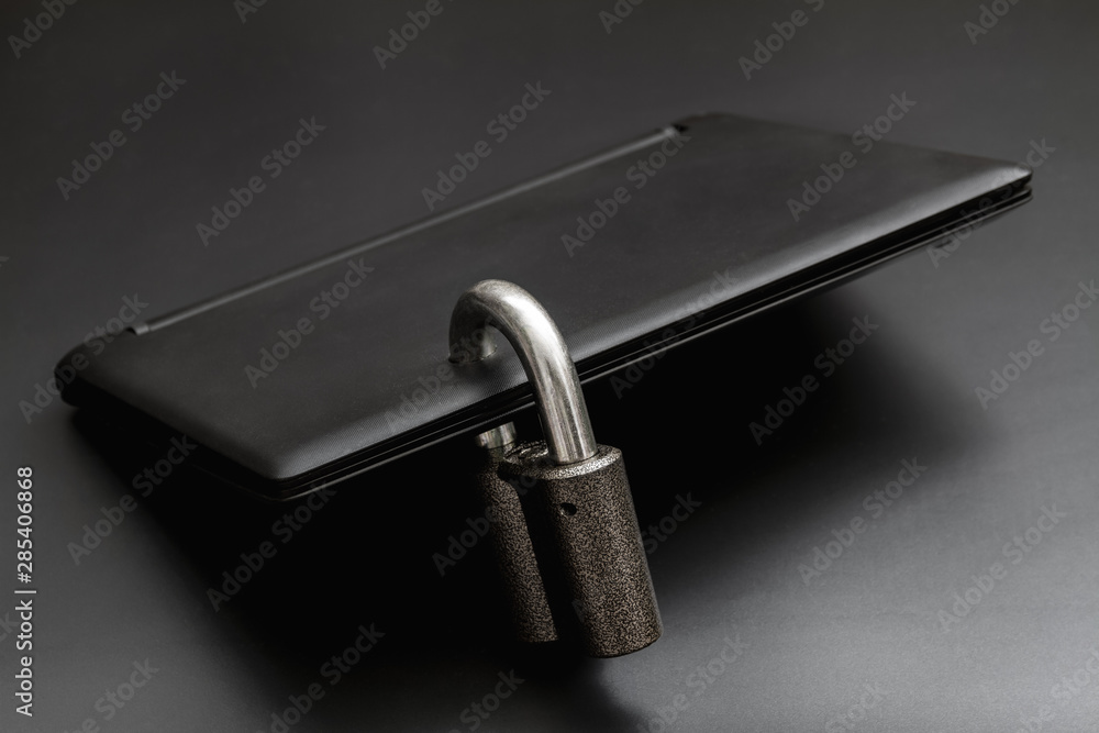 Locked steel padlock in a drilled hole of the black laptop on dark background. Concept of protecting personal data on a computer. A laptop is locked with a lock. Closeup, selective focus Locked steel padlock in a drilled hole of the black laptop on dark background. Concept of protecting personal data on a computer. A laptop is locked with a lock. Closeup, selective focus