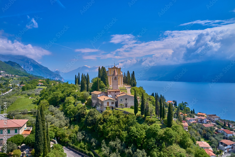 The resort town of Pulciano, located on the shores of Lake Garda. San Michele Arcangelo Church is located on a mountain. Panoramic view of the city, church, Lake Garda, Alps. Aerial view.