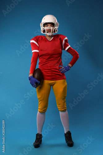 Full-length photo of young American woman football player in white helmet, sportswear with ball in her hand