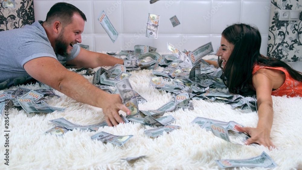 A man and woman share money sitting on a white bed, have fun rushing denbgs and consider them. View from above. Lot of money.Fast money concept
