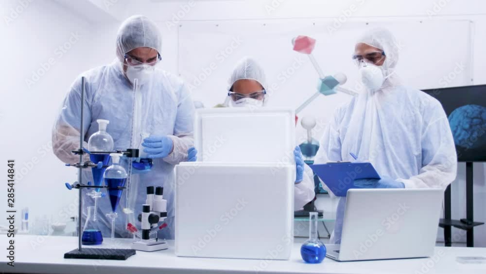 Team of researcher in coverall equipment taking out virus from a refrigerator box. Scientist taking notes. Modern laboratory. Team of researcher in coverall equipment taking out virus from a refrigerator box. Scientist taking notes. Modern laboratory.