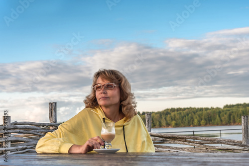 Portrait of a mature woman with a cup of coffee in a summer cafe on the background of the river and forest.