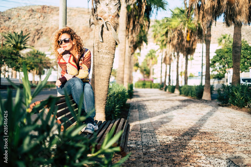 alone and sitting woman in a park with a lot of palms on the background with thoughts and she's thinking about something or looking at the street - casual lifestyle outdoor leisure actiity photo