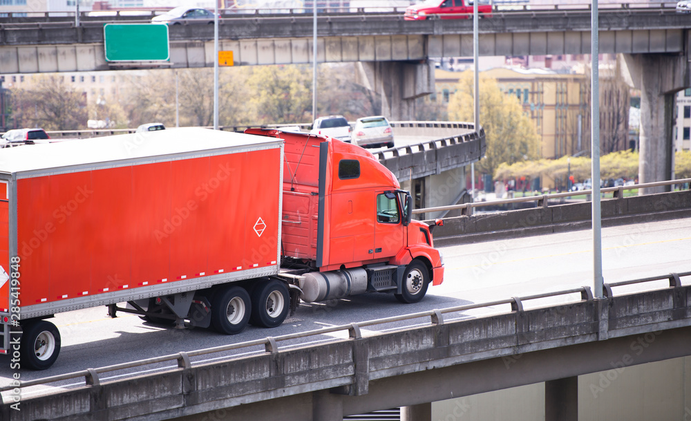 Orange big rig semi truck transporting cargo in dry van semi trailer running on overpass road in urban city