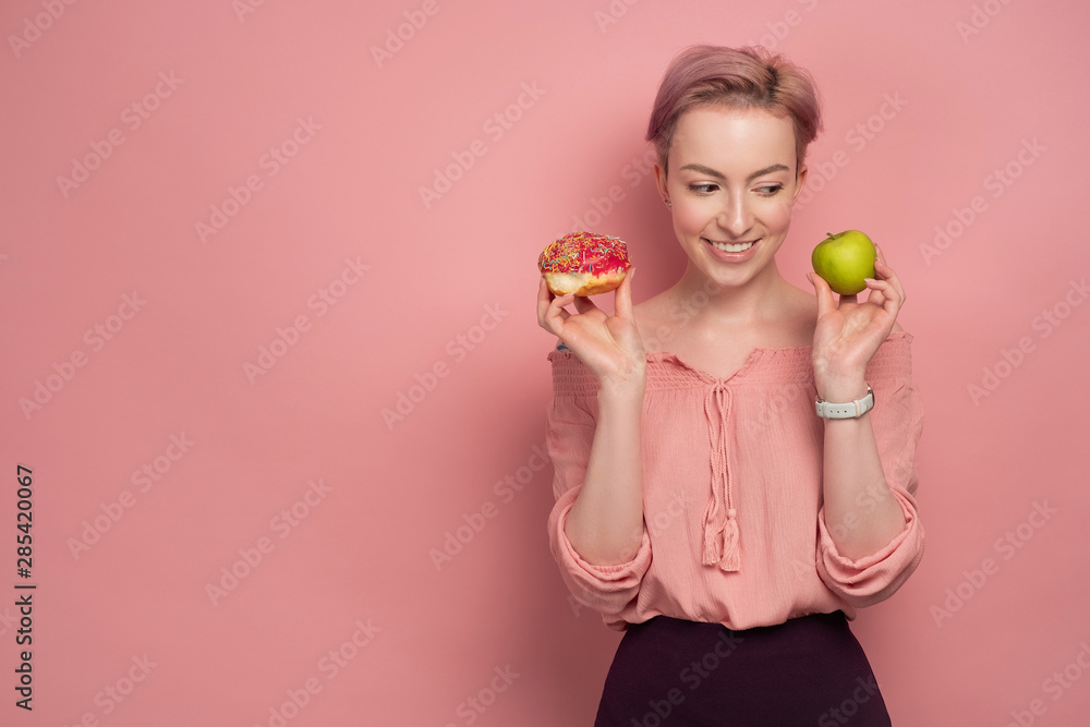 A girl with short pink hair, holds a donut and a green apple near her face, and looks at the apple, smiling, on a pink background. A girl with short pink hair, holds a donut and a green apple near her face, and looks at the apple, smiling, on a pink background.