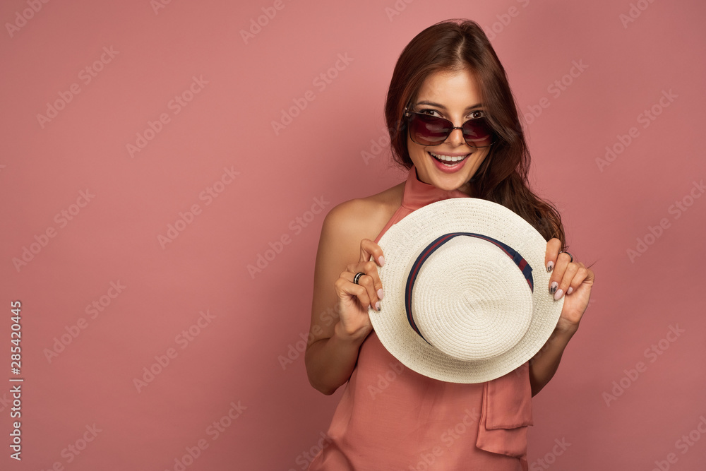 A dark-haired girl in a pink dress and sunglasses looks joyfully into the frame, hiding behind a straw hat on a pink background A dark-haired girl in a pink dress and sunglasses looks joyfully into the frame, hiding behind a straw hat on a pink background