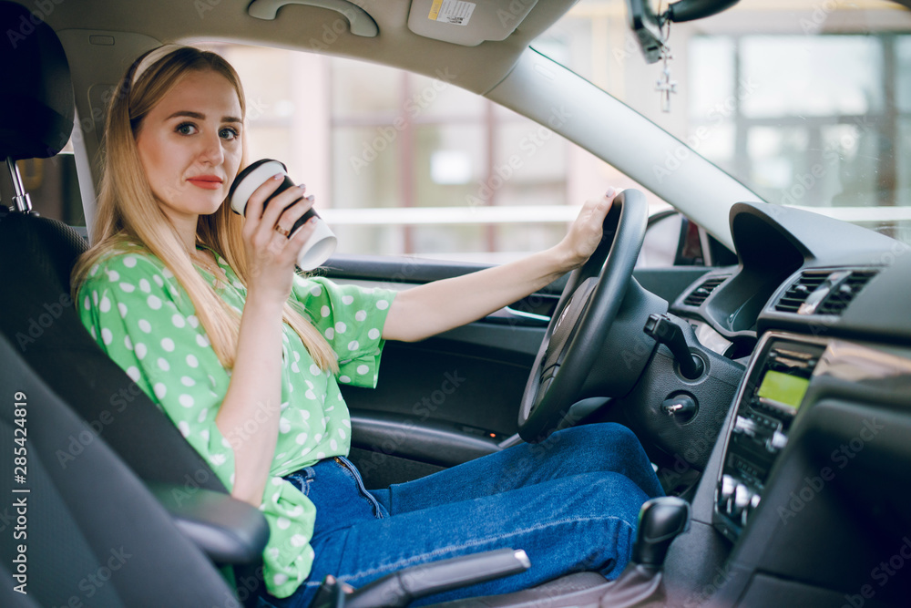 Beautiful blonde girl sitting in the car driving listening to music and drinking coffee