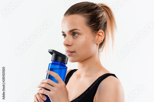 Young sporty blond woman in a black sportswear with smart watches drinks water after workout standing over white background.