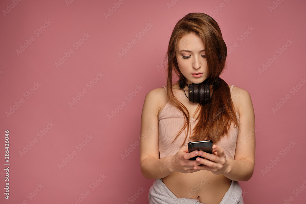 Red-haired girl on a pink background stands in jeans and a top with headphones around her neck looking at the phone in her hands. Red-haired girl on a pink background stands in jeans and a top with headphones around her neck looking at the phone in her hands.