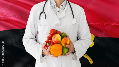 Angolan Doctor is holding fruits and vegetables in hands with Angola flag background. National healthcare concept, medical theme.