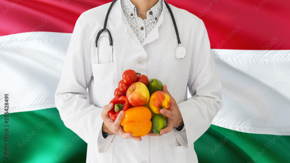 Doctor is holding fruits and vegetables in hands with Hungary flag background. National healthcare concept, medical theme. Doctor is holding fruits and vegetables in hands with Hungary flag background. National healthcare concept, medical theme.