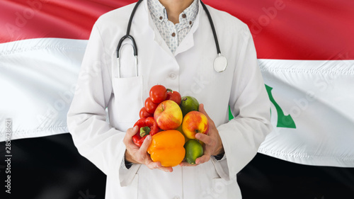 Doctor is holding fruits and vegetables in hands with Iraq flag background. National healthcare concept, medical theme.
