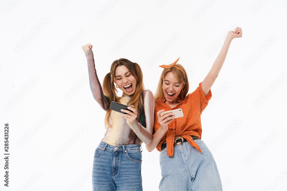 Photo of two excited girls wearing colorful clothes rejoicing while holding smartphones together Photo of two excited girls wearing colorful clothes rejoicing while holding smartphones together