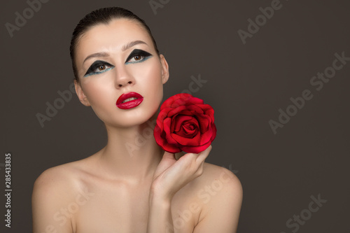 Portrait of a young beautiful woman with bright makeup and red lipstick. Holding a red rose in hands on a dark background. Copy space